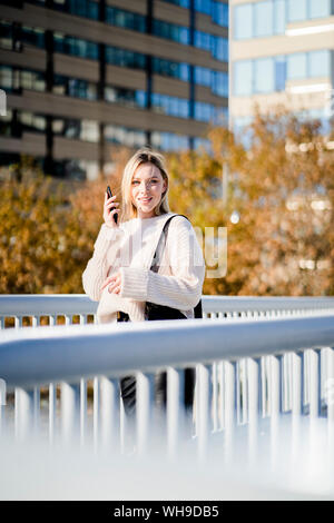 Portrait of smiling young woman with smartphone debout sur la passerelle à l'automne Banque D'Images