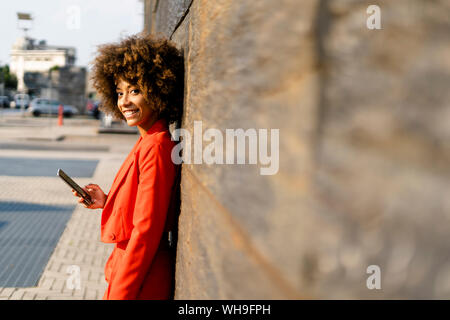 Portrait of smiling young woman with cell phone wearing fashionable tailleur-pantalon rouge leaning against wall Banque D'Images