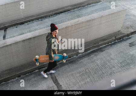 Jeune femme élégante avec skateboard et cell phone marche sur parking Banque D'Images