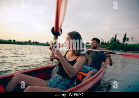 Certain young caucasian couple kayak sur rivière avec coucher du soleil dans l'arrière-plan. Avoir du plaisir dans l'activité de loisirs. Romantique et heureux homme et femme sur le kayak. Le sport, les relations concept. Banque D'Images