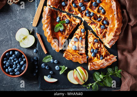 Close-up composé de délicieuses tranches de pomme Tarte aux bleuets sur un plateau en ardoise noire sur une table en béton avec des ingrédients à l'arrière-plan, horizontal vie Banque D'Images