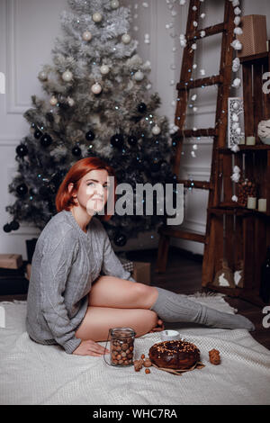 L'intérieur de la mode photo de belle jeune femme aux cheveux rouges et sourire charmeur, tricoté confortable porte cardigan, posant à côté d'arbre de Noël et la présentation Banque D'Images