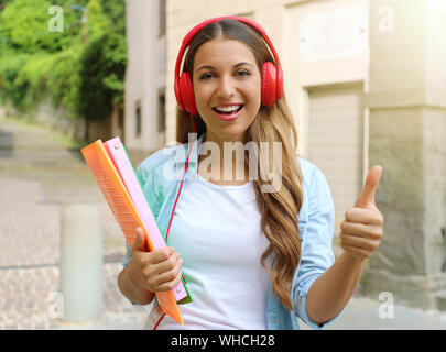 Happy student girl with headphones thumb up donne à l'extérieur. Jeune femme faisant programme d'échange scolaire. Banque D'Images