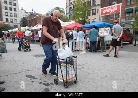 Un homme âgé portant des écouteurs promenades à travers l'Union square Green Market poussant un plein panier. À Manhattan, New York City. Banque D'Images