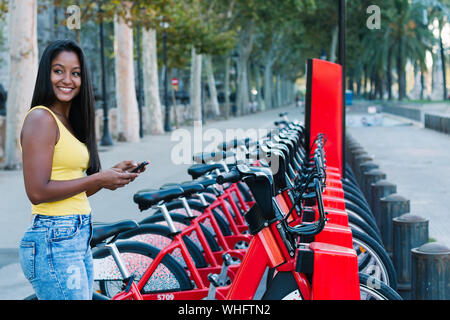 Belle jeune femme de contrôler son téléphone portable tout en louant un vélo dans une ville Banque D'Images