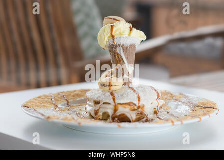 De délicieuses crêpes à la banane et glace à la vanille, sirop sucré, macarons, copeaux de noix de coco sur la table en bois blanc, fond restaurant floue. Banque D'Images