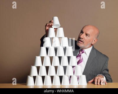 Middle aged businessman monter en pyramide de tasses sur la table contre fond brun Banque D'Images