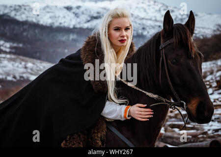 Belle jeune blonde sur un corbeau. Viking femme avec un cheval noir sur fond de montagnes Banque D'Images