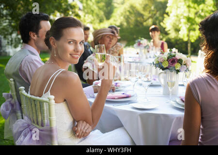 Portrait of beautiful bride holding champagne flute alors qu'il était assis à table avec des invités de mariage Banque D'Images