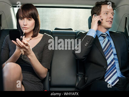 Young businesswoman appliquant un brillant à lèvres et businessman using cell phone in car Banque D'Images