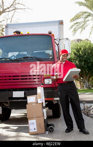 Young delivery man à la liste à la livraison sur le presse-papiers avec chariot en arrière-plan Banque D'Images