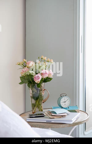 Roses dans un vase sur la table de chevet avec livres et réveil dans la chambre Banque D'Images