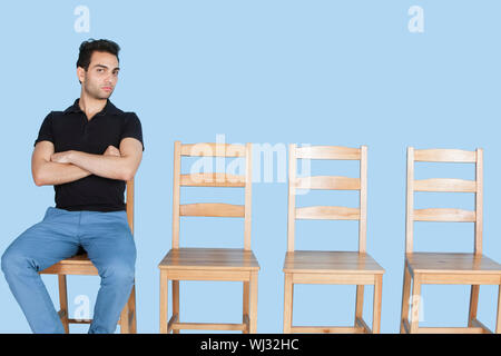 Portrait d'un jeune homme assis à côté de chaises en bois vides sur fond bleu Banque D'Images