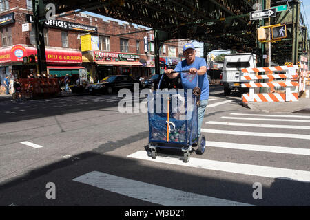 Un homme dans un tee shirt Superman traverser une rue sous le métro surélevé en poussant un panier. Dans la région de Jackson Heights, Queens, New York. Banque D'Images