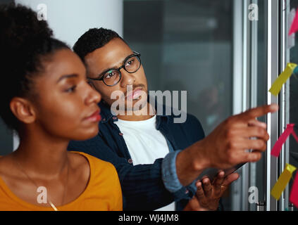 Businesswoman looking at business homme pointant son doigt sur le post-it collé sur le verre Banque D'Images