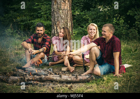 Deux couples camping dans les bois. Saucisses à frire les gars sur le feu. Smiling friends sitting autour de feu de camp le soir Banque D'Images