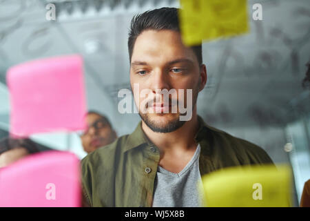 Portrait of a young businessman in casualwear debout derrière le verre transparent en regardant les notes au bureau Banque D'Images