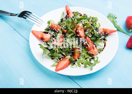 Régime alimentaire sain salade de roquette biologique, les fraises et le sésame avec glaçure balsamique en plaque blanche sur un fond de bois bleu pastel, vue du dessus, de numérisation Banque D'Images