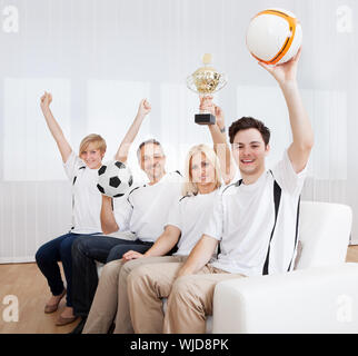 Famille extatique assis sur un canapé célébrer une victoire avec leurs bras levés en liesse brandissant une balle et un trophée Banque D'Images