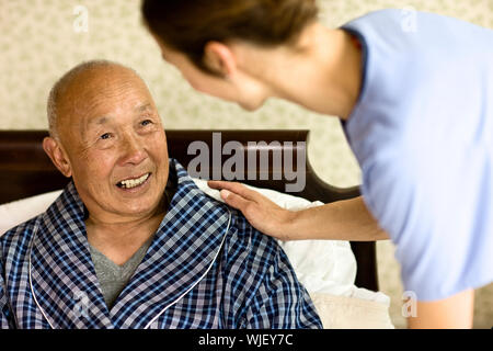 Senior man sitting in bed smiles jusqu'à une infirmière comme elle se penche pour poser une main rassurante sur son épaule. Banque D'Images