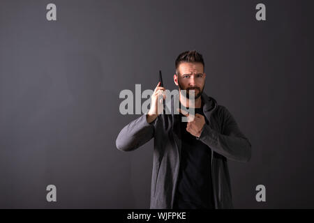 Un beau jeune homme dans un sweat gris en conversation sur son téléphone devant un fond gris foncé dans un studio. Banque D'Images