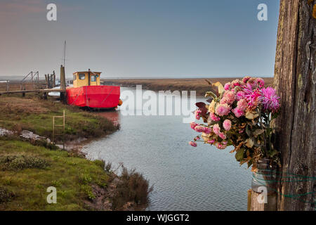 Un bouquet de fleurs rose et lilas attaché à un poteau sur le côté d'un ruisseau des marais salants près de Thornham, Norfolk avec un bateau de pêche dans l'arrière-plan Banque D'Images