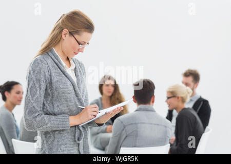 Femme sérieuse thérapeute à écrire des notes avec la thérapie de groupe en session en arrière-plan Banque D'Images