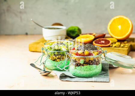 Deux semences chia pudding avec le thé vert matcha, kiwi et granola, orange, dans les verres sur fond de béton rose. Petit déjeuner sain. Banque D'Images