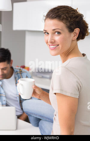 Cheerful woman drinking coffee alors que des partenaires utilise ordinateur portable à la maison dans la cuisine Banque D'Images