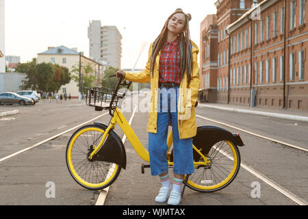 Jeune femme portant manteau de couleur jaune et des tresses, équitation, vélo en ville. Banque D'Images