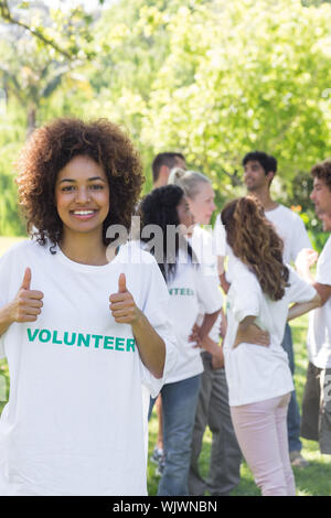 Confident female volunteer gesturing Thumbs up avec des amis en arrière-plan disucssing Banque D'Images