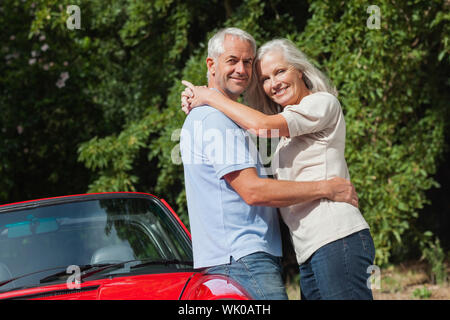 Smiling mature couple hugging against their red cabriolet Banque D'Images