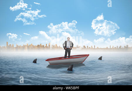 Smiling businessman with hands on hips contre les requins tournant autour d'une petite embarcation à la mer Banque D'Images