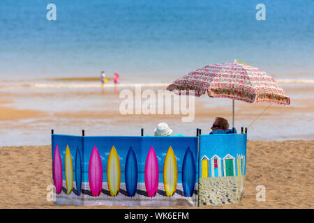 Un couple ayant une journée sur la plage de Colwyn Bay sous un parasol et derrière un brise-vent de planche à voile, au nord du pays de Galles Banque D'Images