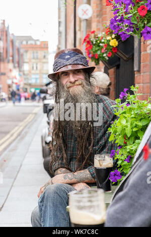 Belfast, en Irlande du Nord, ROYAUME UNI - 30 juillet 2019 : Portrait d'un homme mûr avec une longue barbe et portant un chapeau assis dans la rue ayant une bière à Belfast Banque D'Images