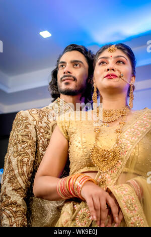 Groom indiens habillés en Sherwani avec bride standing avec superbe tenue glamour et des bijoux avec le maquillage et tenez chaque main dans une salle de banquet. Banque D'Images