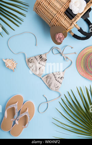 Fond d'été avec des feuilles de palmier, fashion hat, bikini, tongs, sac de plage de paille sur un fond bleu pastel, voyage et vacances concept, Banque D'Images