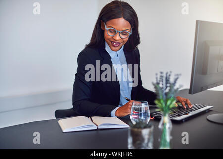 Smiling young African American businesswoman travaillant sur un ordinateur et lire des notes alors qu'il était assis à son bureau dans un bureau Banque D'Images