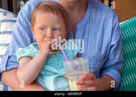 En plein air de l'alimentation avec des bébés. Petite fille est assise sur les genoux des mères. La Nutrition du bébé. Une saine alimentation solide pour peu de clients. La fraîcheur des boissons de jus de fruits, avec de la paille bébé fille blonde aux yeux bleus Banque D'Images