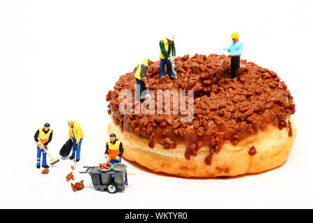 Image conceptuelle d'une figure miniature Ouvriers debout sur un beignet au chocolat faire le trou au centre alors que les nettoyeurs tidy up garnitures tombé Banque D'Images