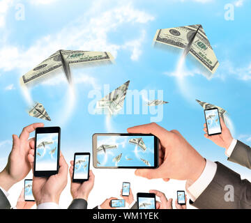 Mains tenant des téléphones intelligents et des vidéos comme flying paper plane de dollars. Ciel avec nuages en arrière-plan Banque D'Images