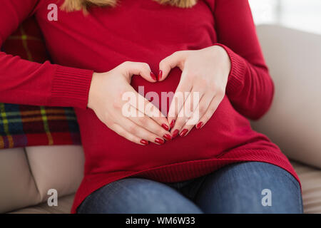 Près d'une femme enceinte faisant coeur signe sur son ventre à la maison dans le salon Banque D'Images