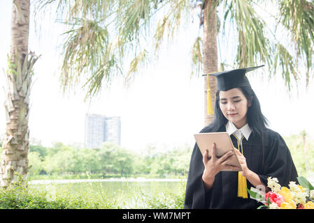 Belle femme de son diplôme à l'aide de tablet pc. Femme asiatique diplômés student wearing graduation hat et une blouse ,le fond est la nature à l'université. Banque D'Images