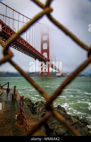 Le célèbre Golden Gate Bridge vue à travers une clôture à mailles de chaîne rouillée sur une journée d'été à portée de roulement de brouillard à San Francisco, Californie Banque D'Images