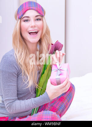 Portrait of happy smiling girl sitting on la lit dans pajama et profiter de la Saint-Valentin romantique présente, recevoir des fleurs et petit cadeau fort, l'amour Banque D'Images