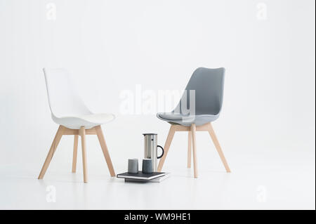 Deux chaises modernes avec une portion de café dans un ballon avec deux mugs reposant sur des livres reliés sur le sol entre les deux, sur fond blanc Banque D'Images