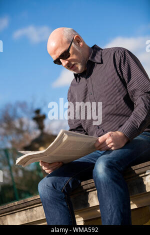 Middle-aged homme chauve assis au soleil en lisant un journal sur un mur de pierre dans un environnement urbain avec sa veste plié à ses côtés Banque D'Images