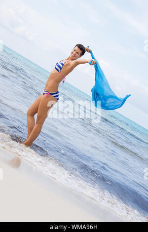 Belle jeune femme insouciante dans un bikini en marche et sautant sur une plage avec un joli foulard bleu turquoise en flottant dans l'air derrière elle alors qu'elle Banque D'Images