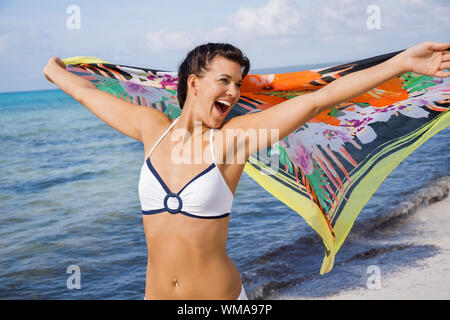 Femme vivace rire dans un bikini au bord de la mer, tenant une écharpe à motifs colorés dans sa main tendue à flottent dans la brise contre un Banque D'Images