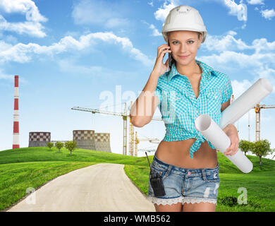 Woman in hard hat, debout sur la route entre les collines vertes, holding Dimensions rouleaux, appelant au téléphone, looking at camera, souriant. Grues et fumer-s Banque D'Images